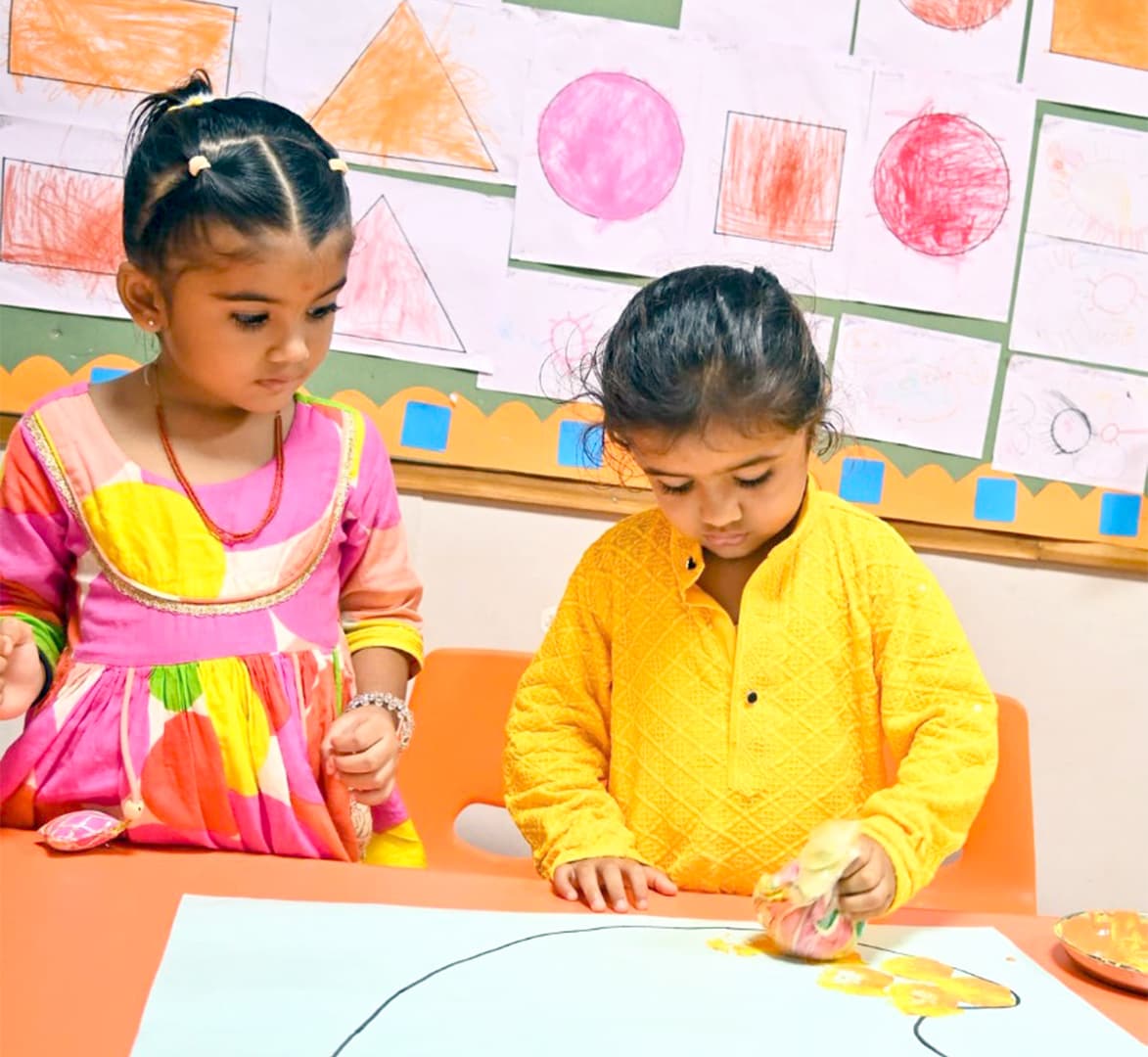 Two young students painting on a large sheet of paper below a wall of their artwork