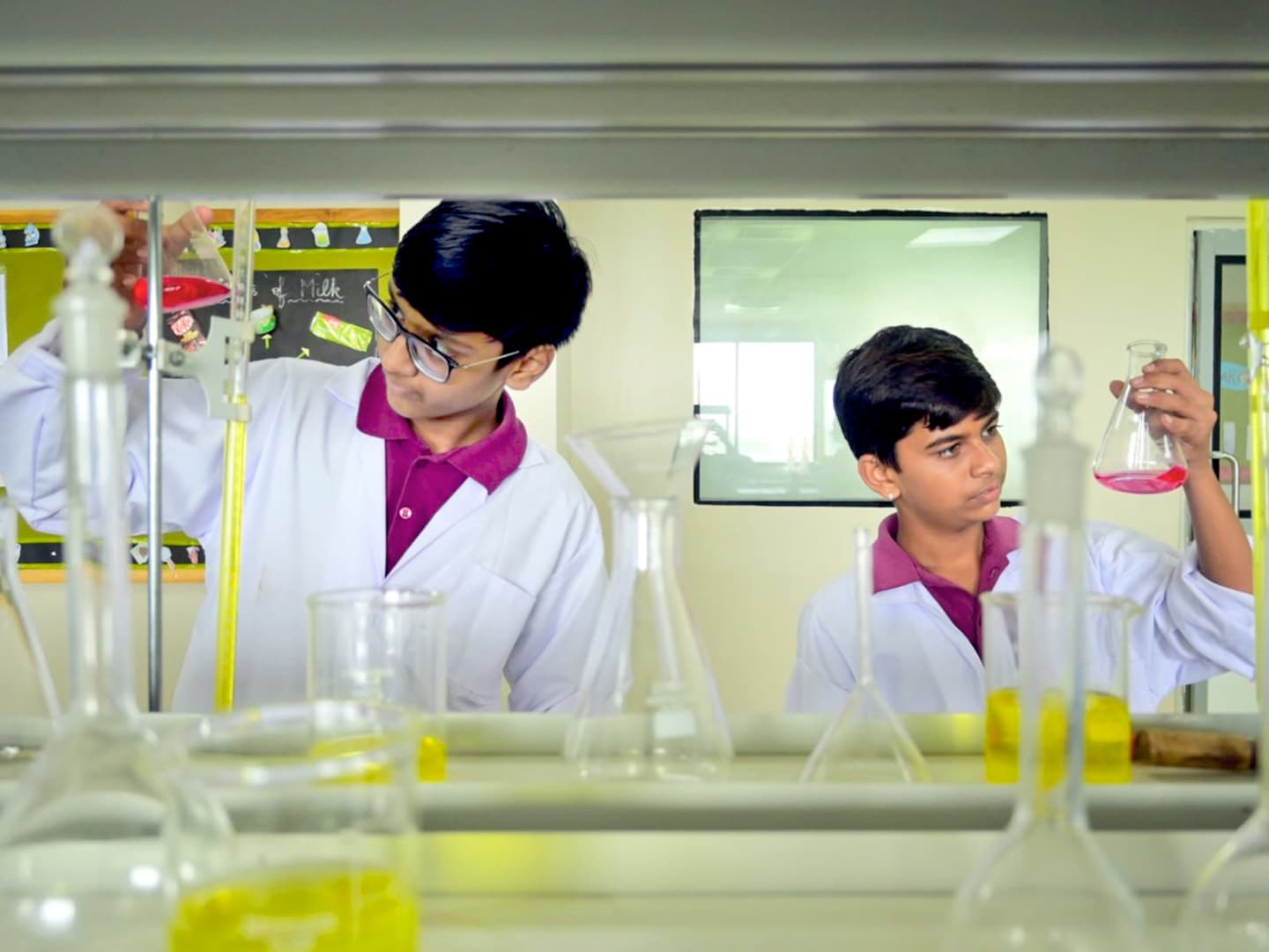 Two students in lab coats measuring liquids into flasks in the school chemistry lab